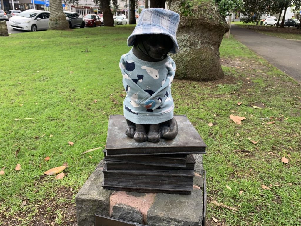 Statue of Benjamin the library cat. It sits on a pile of books. The statue is given outfits - and today he's wearing a blue and white plaid hat and a sweat shirt with whales and seals on.
