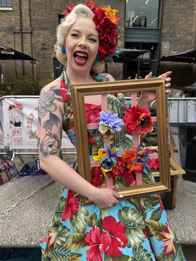 Self Raising Flowers stall at Kings Cross Classic Car Boot Sale. Woman stands holding flower headphones. She is dressed in a 1940s Dior Style dress with flowers in her hair.