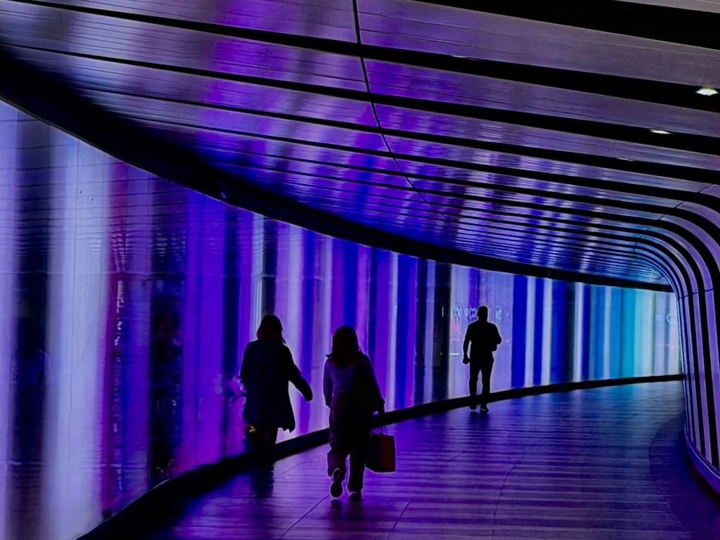 People walk along the coloured tunnel in Kings Cross, London. Purple lights shine along the walls