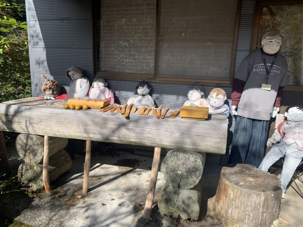 Scarecrows lined up behind a wooden bench displayed carved items like shoehorns for sale at Scarecrow Village