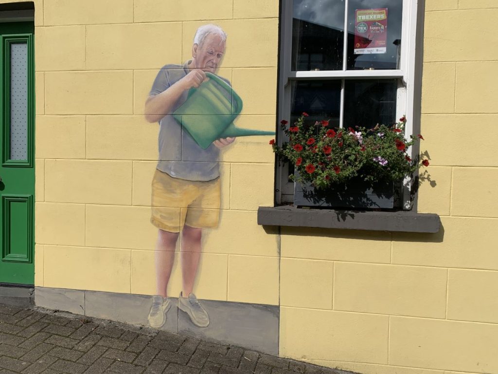 Mural of man dressed in a grey tee shirt and yellow shirts with a green watering can. the can leans into some real flowers on a windowsill of a house in Letterkenny, Ireland